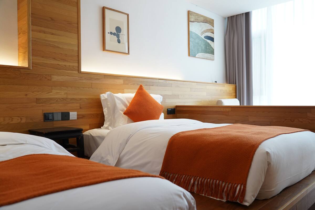 A hotel room with two beds, wooden headboard, white bedding, orange throw pillows, and artwork on the walls.