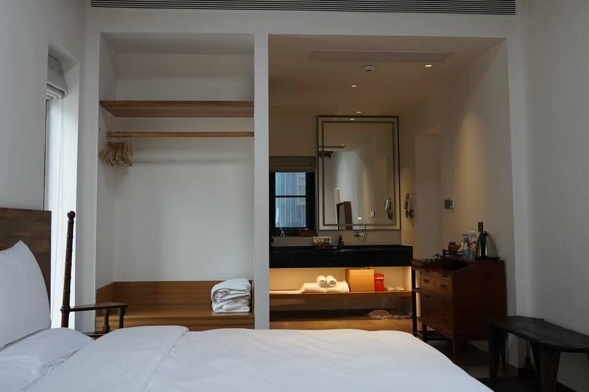 A bedroom with a large bed, wooden headboard, a bench, and a view into a bathroom with a mirror and sink.