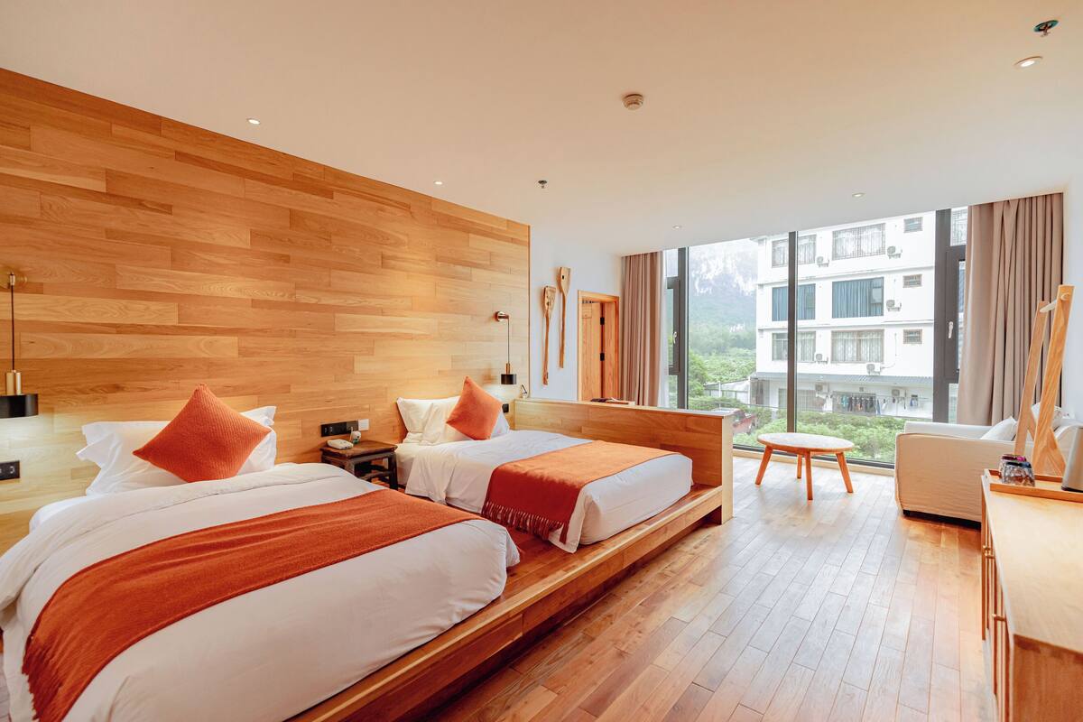 A modern hotel room with two beds, wooden flooring, a large window with a view of buildings, and a small table.