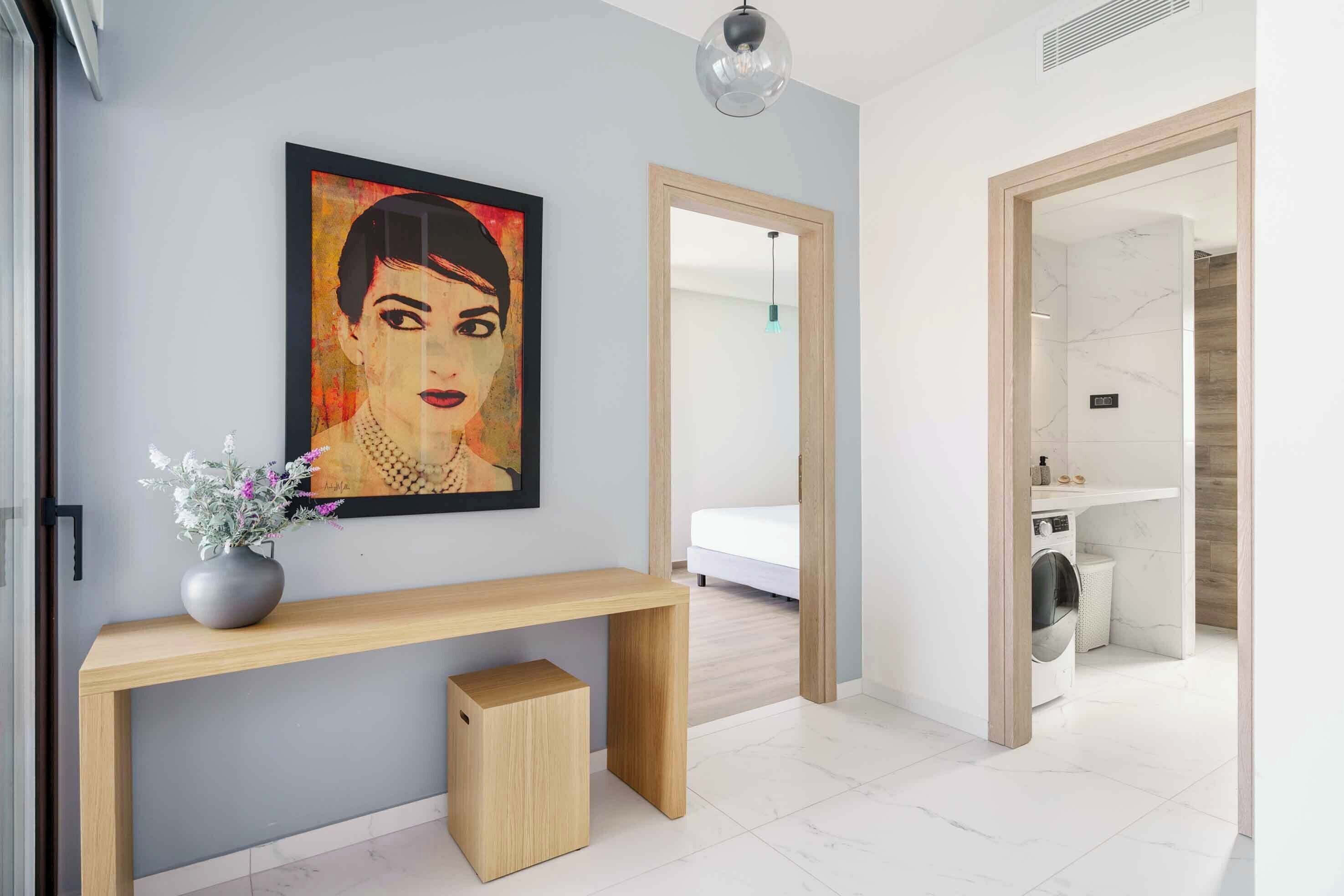 A modern interior with a wooden console table, a vase with flowers, a painting, and a laundry area with a washing machine.