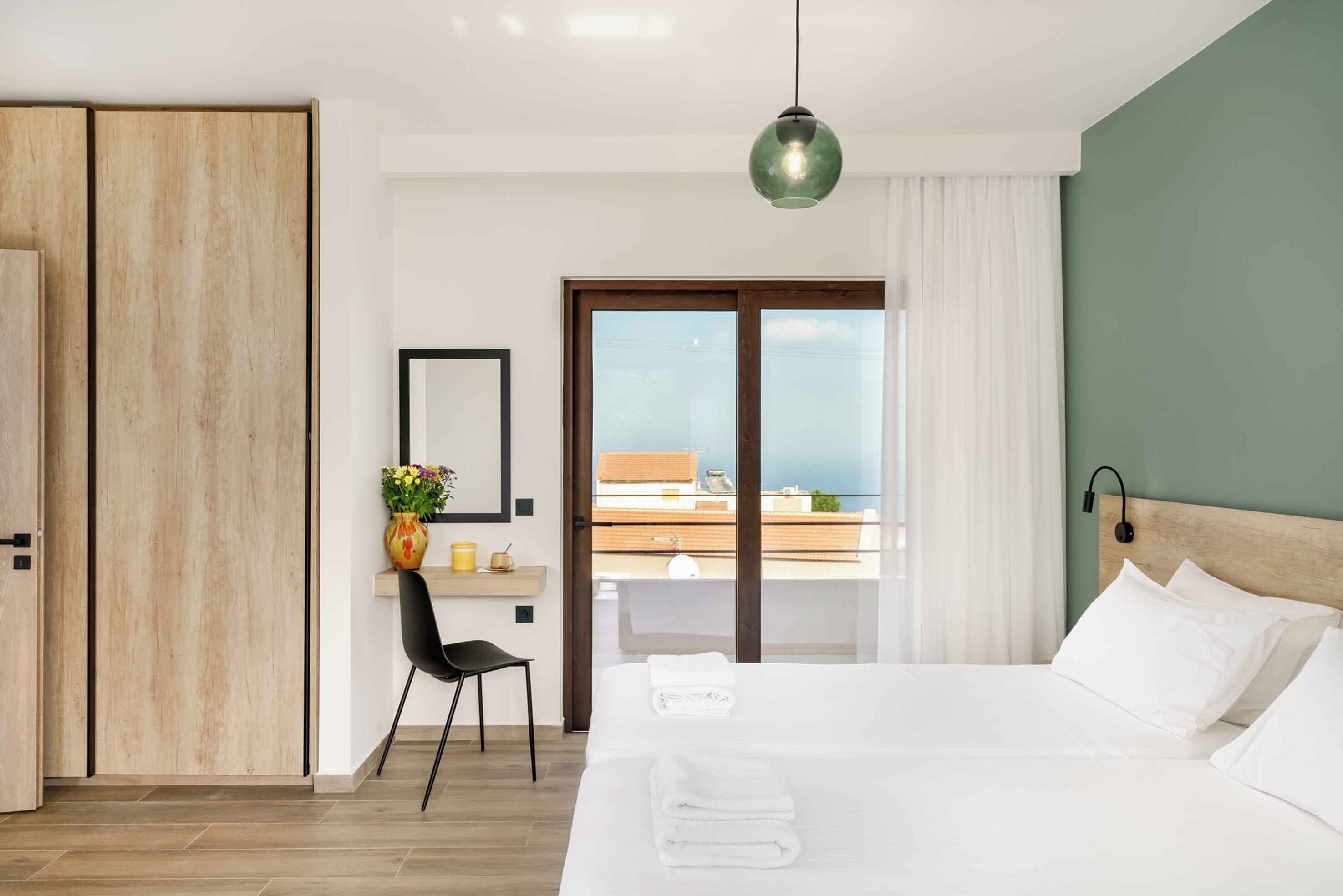 Une chambre d’hôtel moderne dotée d’un grand lit, d’un bureau avec une chaise, d’une vue sur la mer grâce à une porte coulissante et d’un luminaire suspendu.