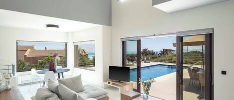 A modern living room with a large sofa, a coffee table, and a TV. There is a sliding glass door leading to a pool area with outdoor furniture.