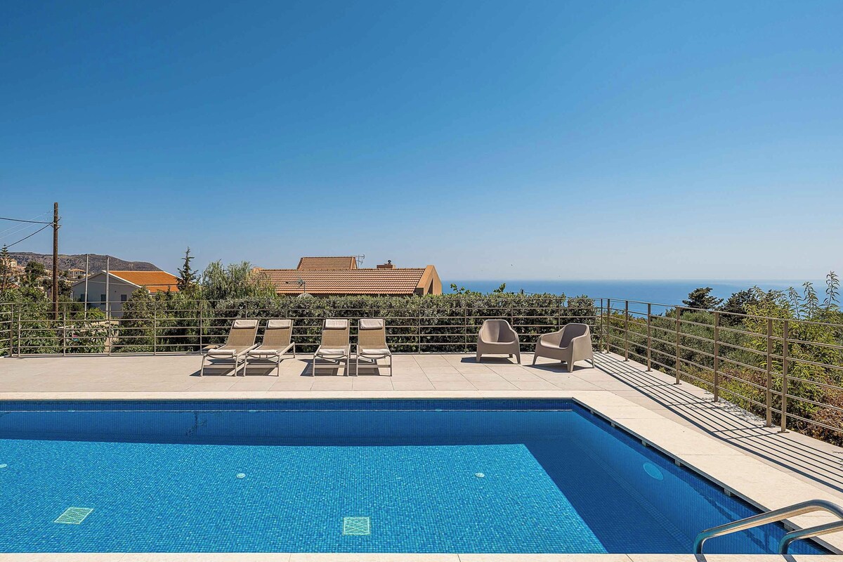 A swimming pool with lounge chairs, a view of the sea, and buildings in the background.