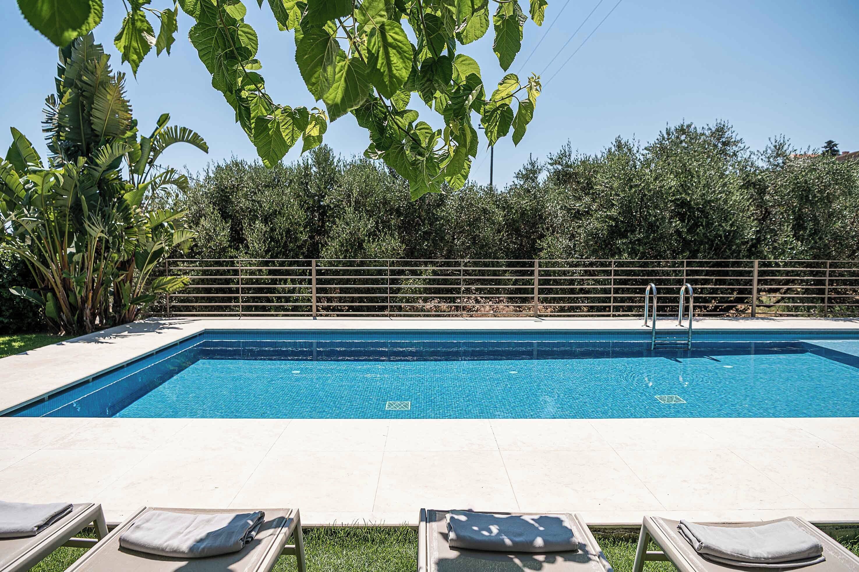 Une piscine rectangulaire, bordée d’une terrasse carrelée, entourée d’une clôture en métal, et des chaises longues à l’avant-plan.