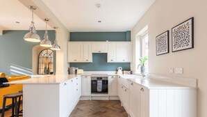 Uma cozinha com armários brancos, piso de madeira em padrão herringbone, iluminação pendente e um sofá amarelo.