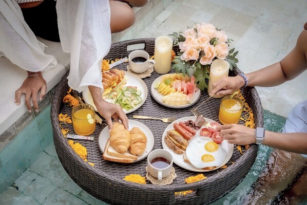 A wicker table with breakfast items, including croissants, eggs, and fruit, surrounded by candles and flowers. - Villa Ubuntu at Bocoa Villas (Canggu)