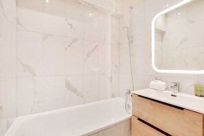 A modern bathroom with a marble-tiled shower wall, a white bathtub, and a wooden vanity with a mirror.