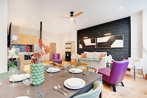 A modern dining area with a wooden table set for four, surrounded by contemporary chairs. A black brick wall and a large abstract painting are visible in the background.