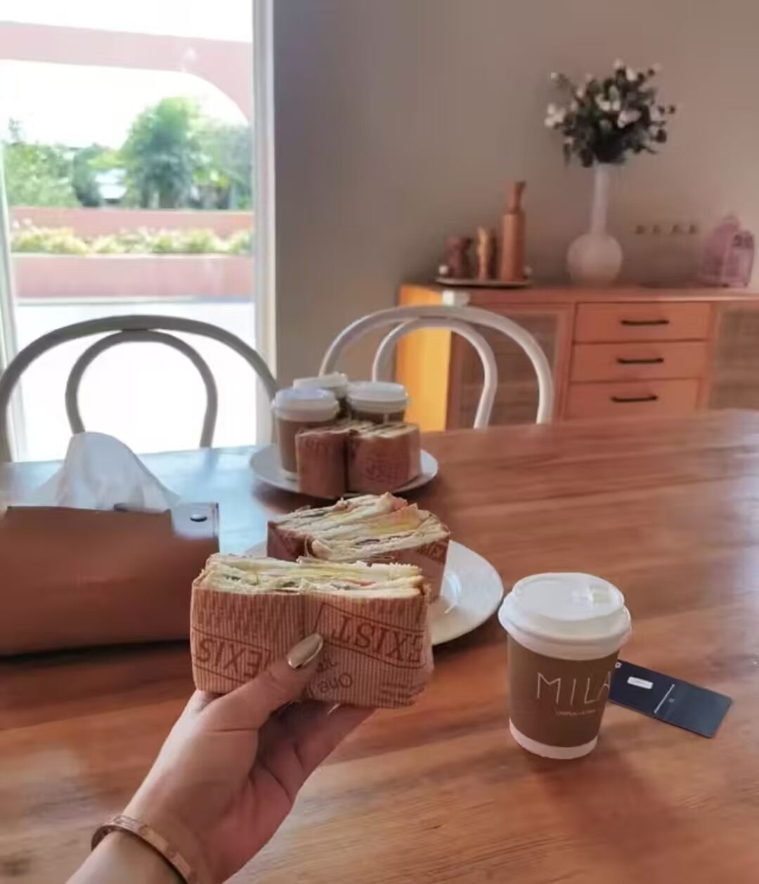 Eine Hand hält ein Sandwich mit sichtbarem Text auf der Verpackung, eine Kaffeetasse mit Deckel und im Hintergrund ein Tablett mit Sandwiches.