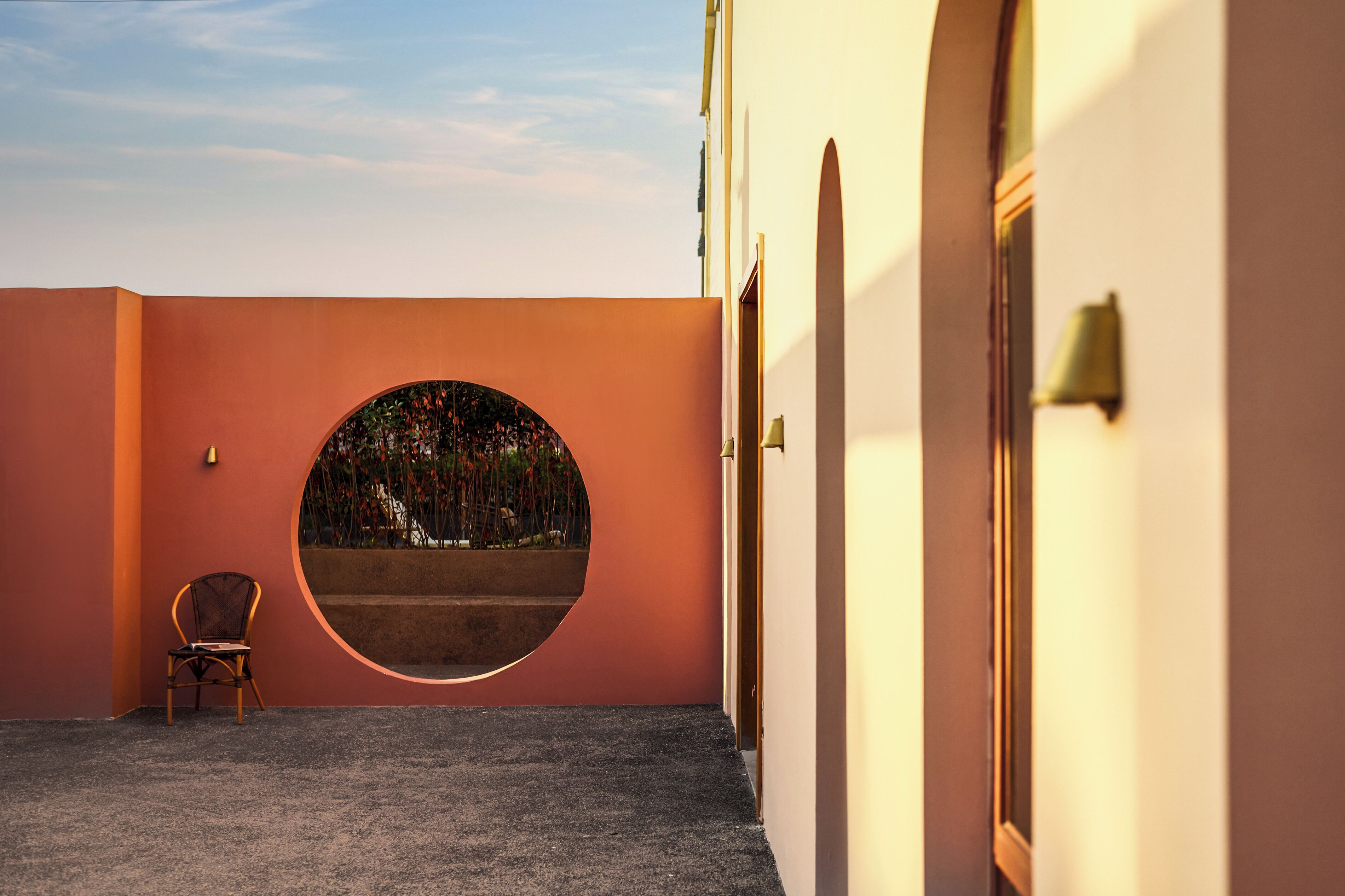 A circular window with a view of trees and a chair in a corridor with a curved wall.