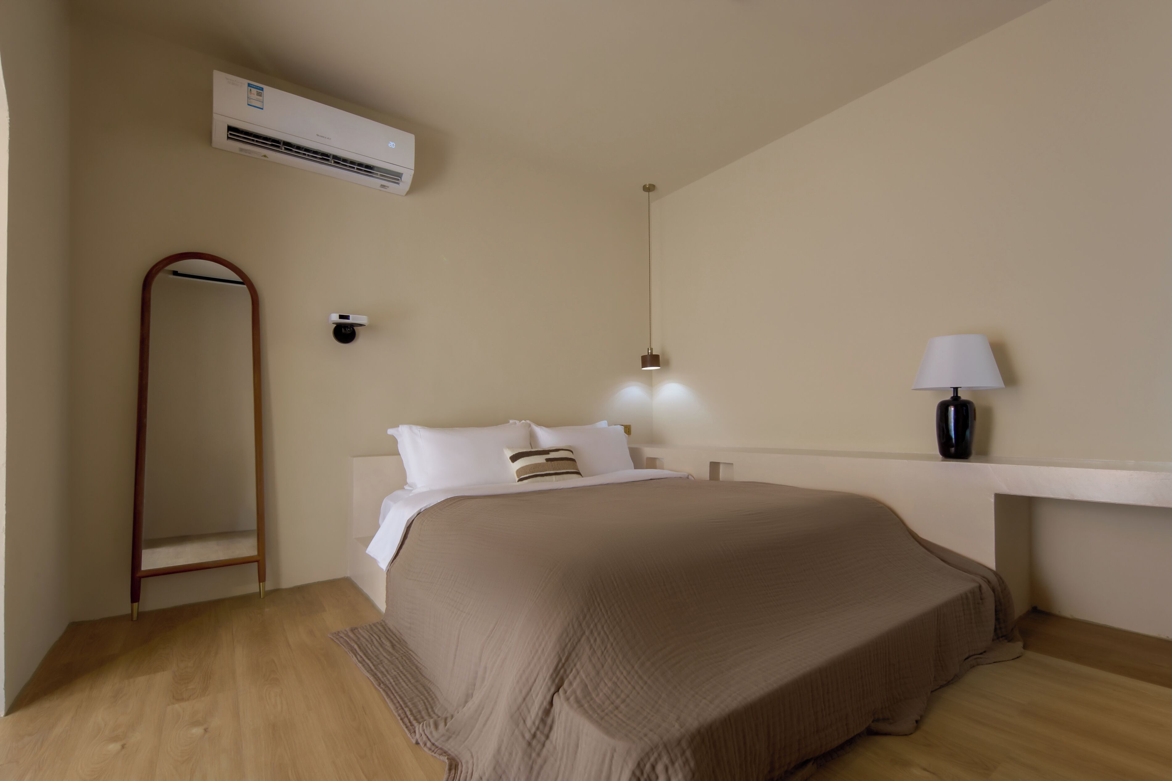 A bedroom with a bed, a bedside table with a lamp, an air conditioning unit, and a mirror.