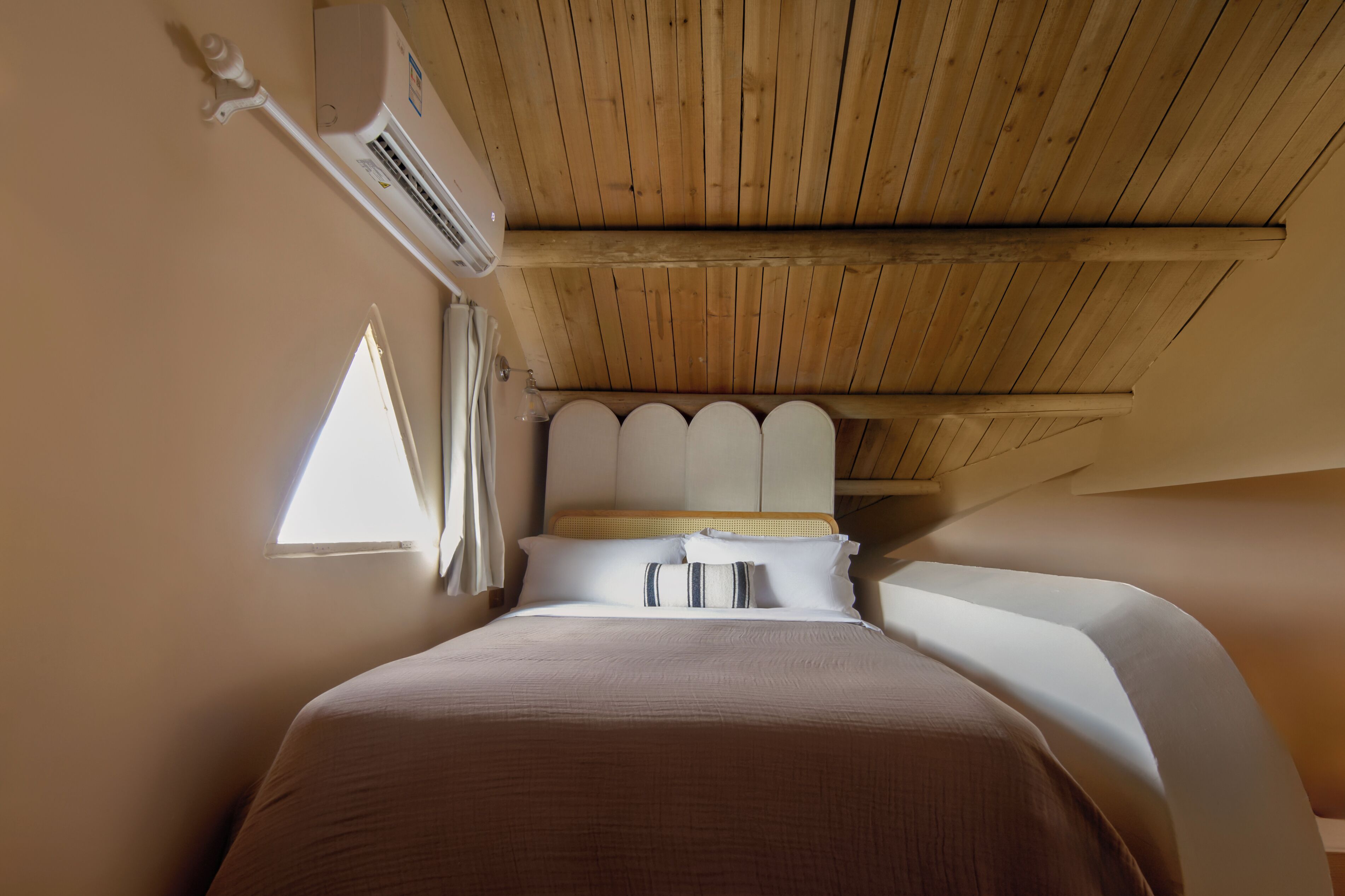 A bedroom with a wooden ceiling, a bed with white bedding, and a window providing natural light.
