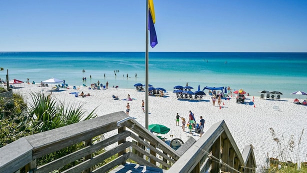 A beach with a flagpole, beach chairs, umbrellas, and a wooden walkway.