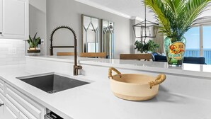 Une cuisine moderne avec un plan de travail blanc, un évier noir et un saladier en bois. On y trouve une plante en pot et un vase décoré sur le thème de la plage.