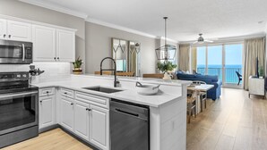 Une cuisine moderne avec des meubles blancs, un four à micro-ondes encastré et un lave-vaisselle. Un coin repas est aménagé avec un canapé bleu et offre une vue sur l’océan grâce à de grandes fenêtres.