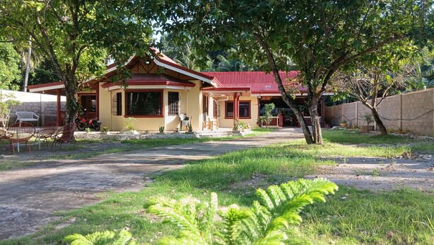 A two-story house with a red roof, a porch with chairs, and a small garden area. - Jovick Homestay (Siquijor)