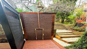 A wooden outdoor shower with a metal rain showerhead, surrounded by a wooden deck and a stone pathway.