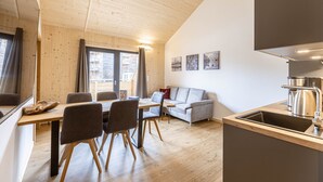 A modern kitchen with wooden flooring, a dining area with a table and chairs, a grey sofa, and a large window with curtains. - Charming Apartment in Oberwolz Near ski Resort (Oberwölz)