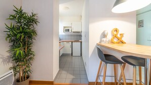 Uma cozinha moderna com armários brancos, micro-ondas e pia. Há uma planta em vaso à esquerda e uma mesa de madeira com duas cadeiras.