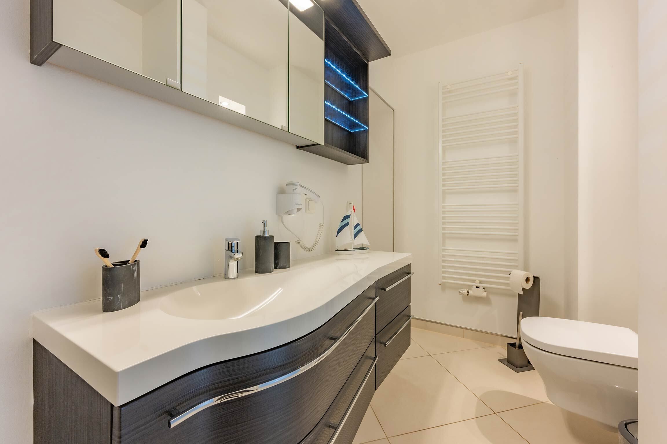 Een moderne badkamer met een witte wasbak, donkere kastjes, een spiegel met blauwe LED-verlichting, een toilet en een raam met blinds.