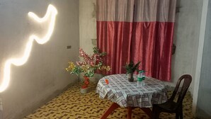 A small room with a patterned floor, a table with a patterned tablecloth, two potted plants, a chair, and a curtain. - Kalzug (Katihar)