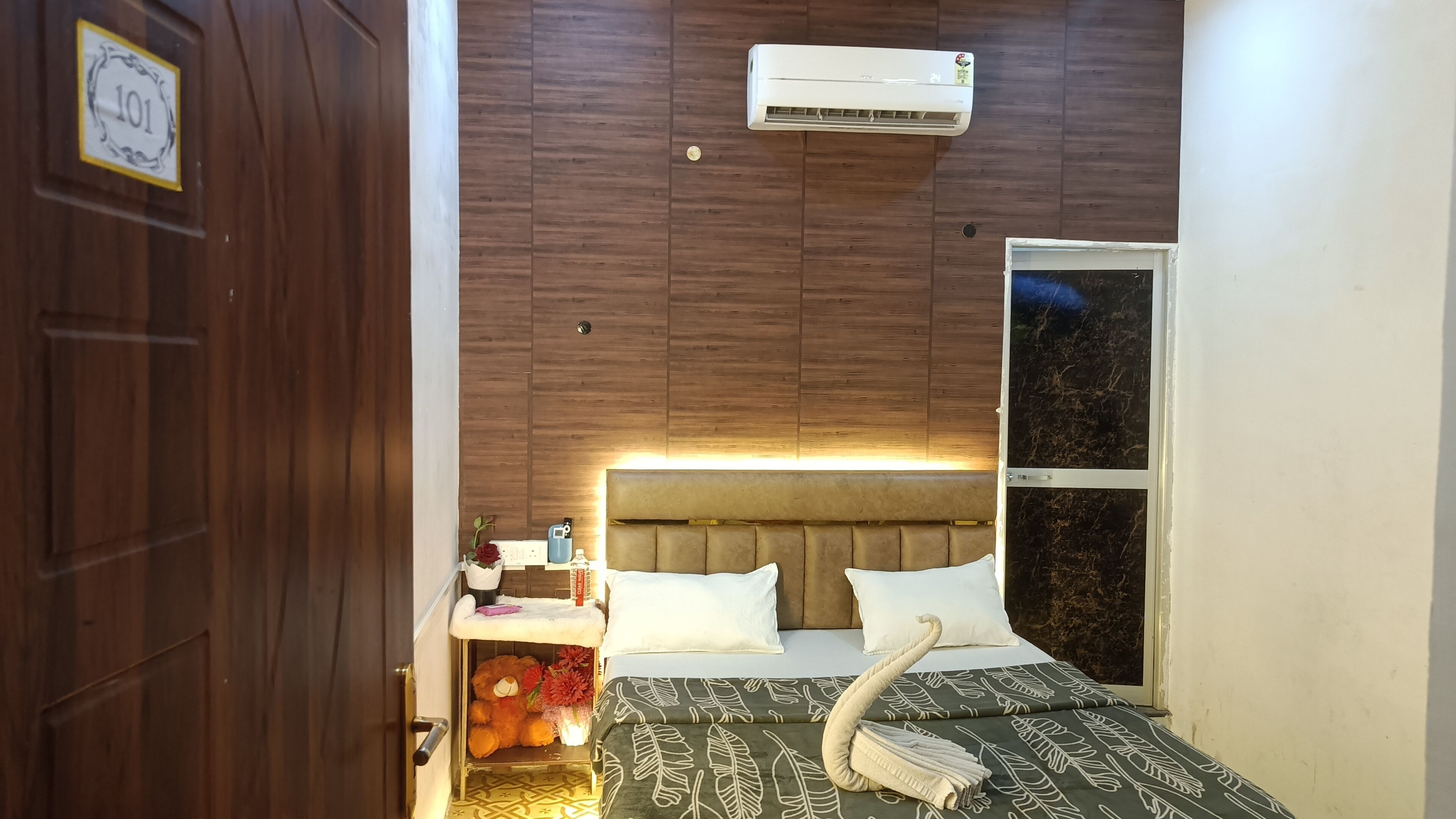 Un dormitorio con cabecera de madera, cama con ropa de cama blanca, mesita de noche con lámpara, un osito de peluche y una ventana con vista al cielo nocturno.