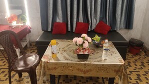 A room with a sofa, red cushions, a table with a floral tablecloth, a chair, and a window with curtains. - Kalzug (Katihar)