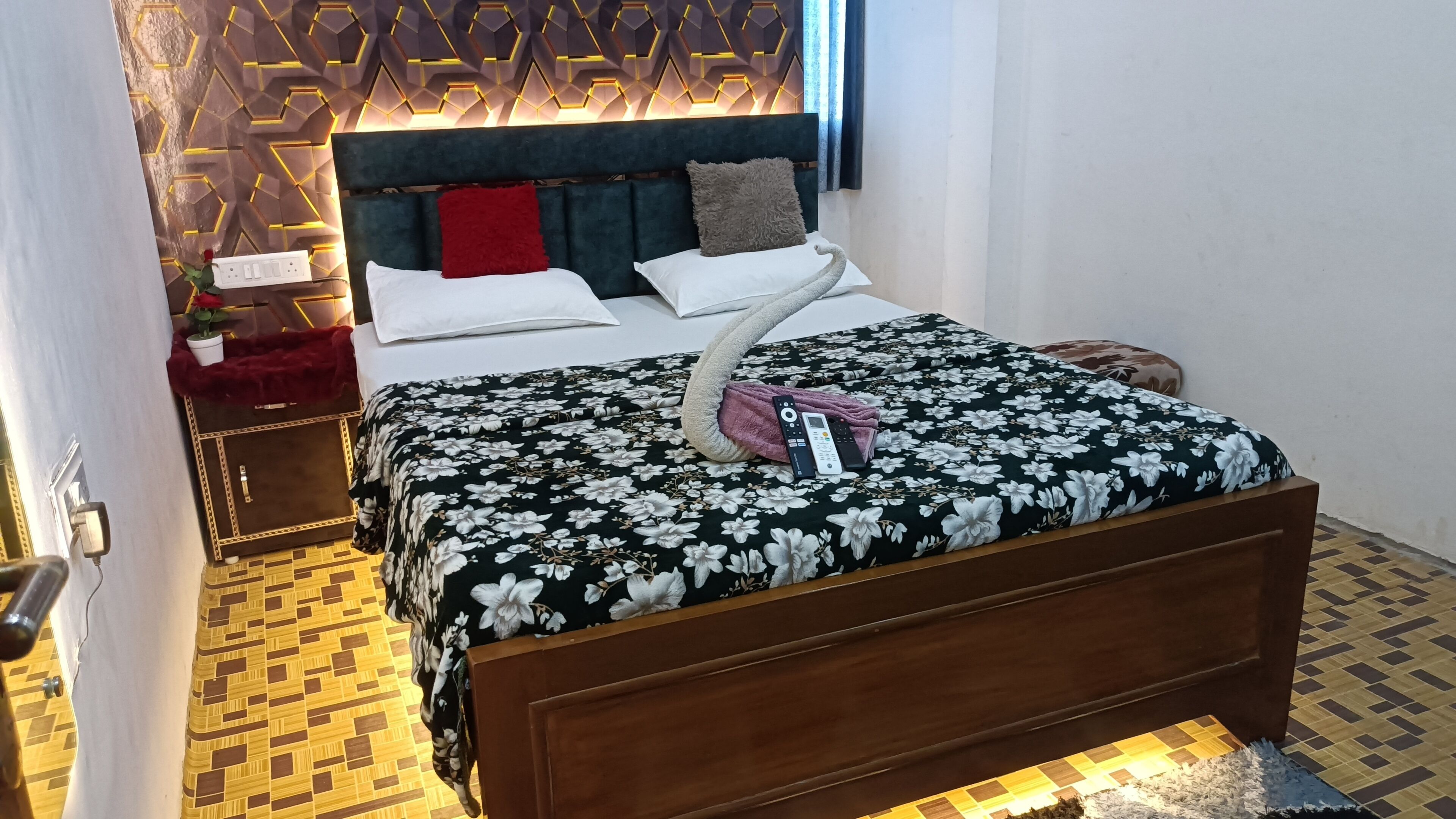 Una cama bien hecha con una colcha floral, un cabecero con diseños geométricos y una mesita de noche con un paño rojo y una planta en maceta.