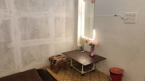 A small, dimly lit room with a single bed, a small stool, a vanity table with a mirror and a vase of flowers, and a red trash bin. - Kalzug (Katihar)