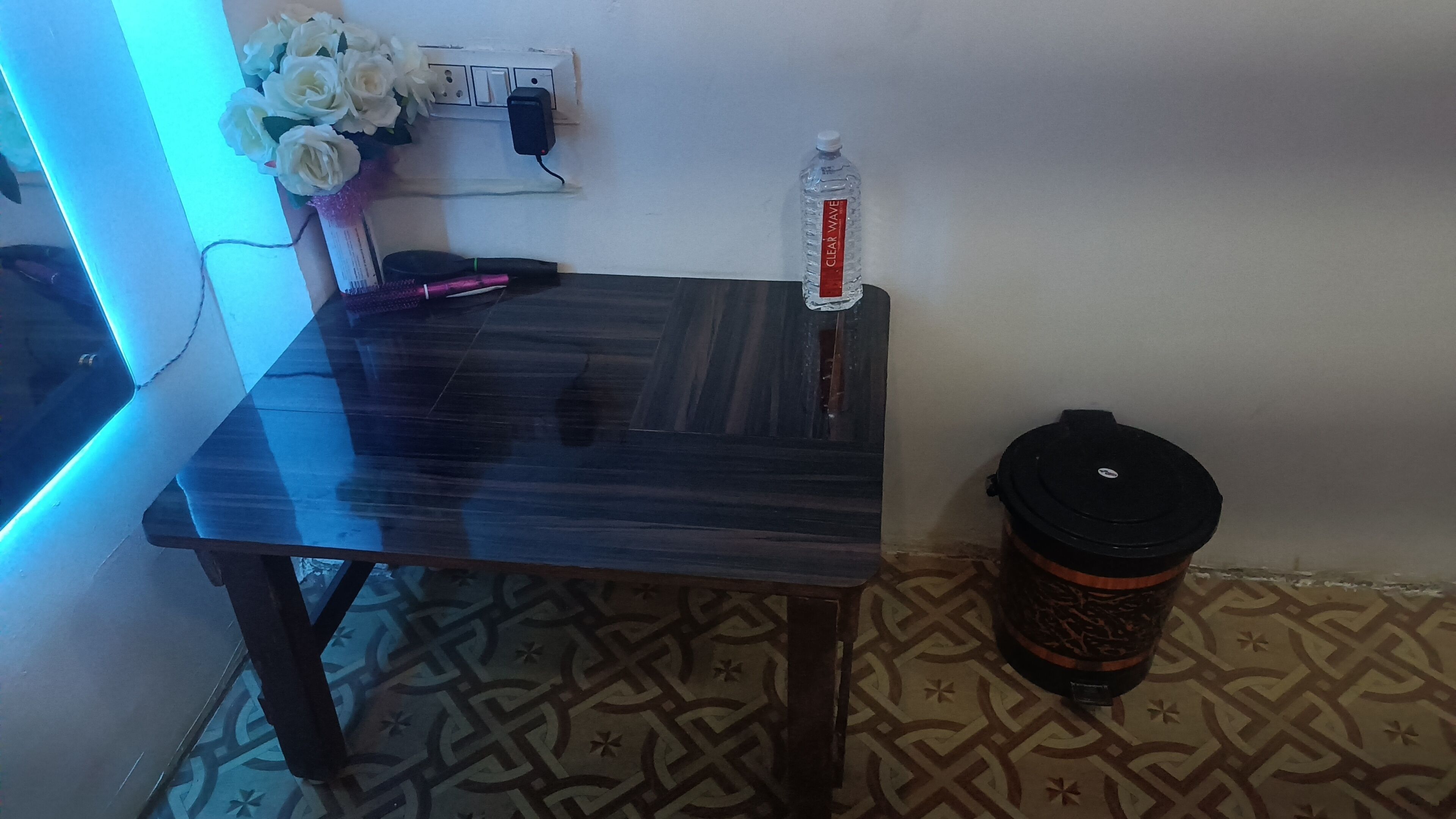 Una habitación con una mesa de madera oscura, una botella de agua, un cesto de basura y un enchufe con un tapón.