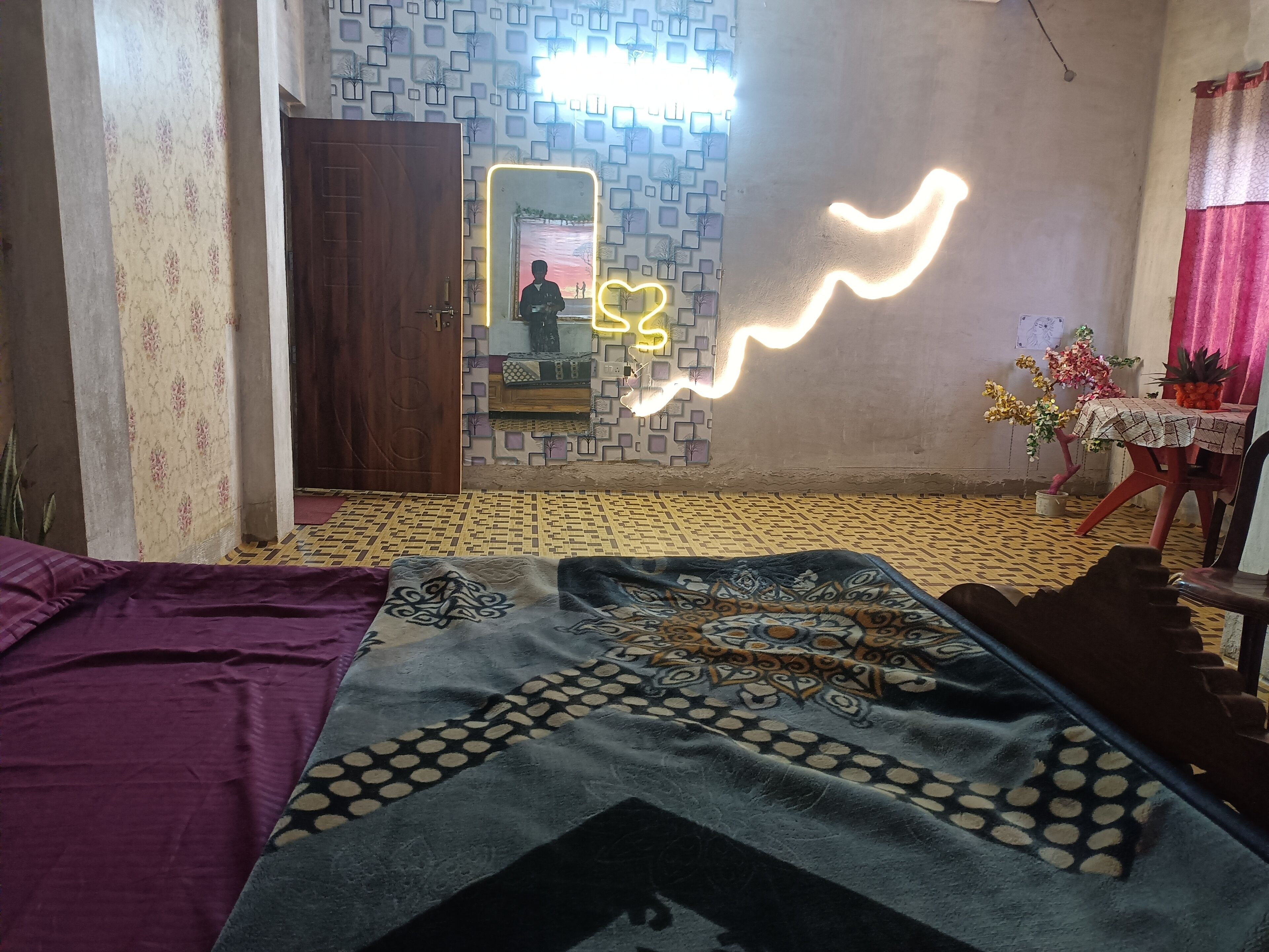 A room with a patterned floor, a bed with a patterned blanket, a mirror with a neon light, a table with a plant, and a wall with a geometric pattern.