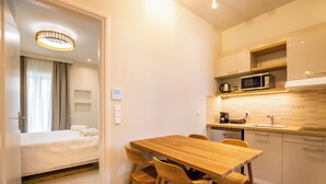 A compact hotel room with a bed, a wooden dining table with chairs, a kitchenette with a microwave, and a ceiling light fixture. - Onyro Villas (Karpathos)