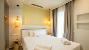 A hotel room with a large bed, two bedside lamps, a wooden side table, a built-in shelf, and a window with curtains. - Onyro Villas (Karpathos)
