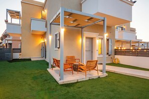 A modern outdoor seating area with a pergola, artificial grass, and a white door. - Onyro Villas (Karpathos)