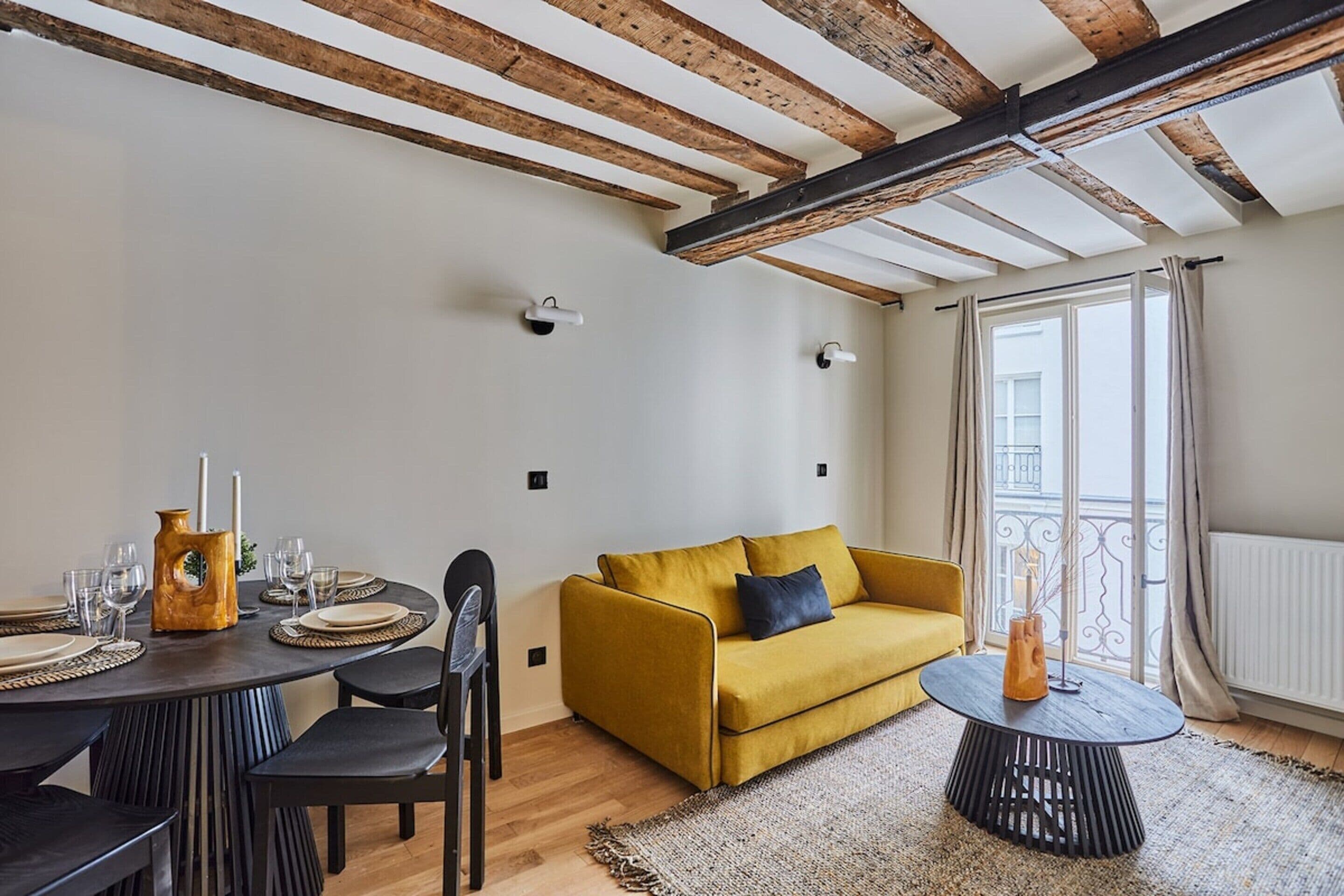 A modern living room with a yellow sofa, wooden beams on the ceiling, a dining table set with plates and glasses, and a large window with curtains.
