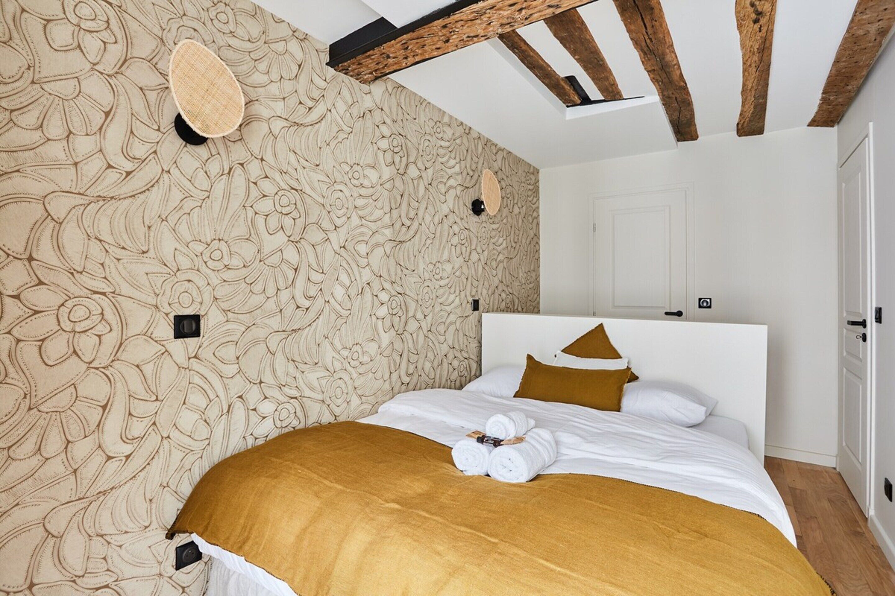 Ein Schlafzimmer mit einer blumengemusterten Wand, einer Holzbalkendecke und einem Bett mit weißer Bettwäsche und einer braunen Tagesdecke.