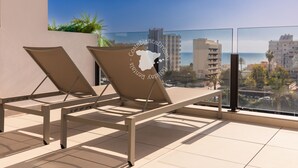 Een balkon met twee ligbedden, een glazen hekwerk en uitzicht op zee en gebouwen.