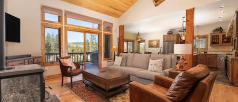 A spacious living room with a fireplace, a large sofa, and a coffee table. There is a kitchen area with wooden cabinets and a dining table.