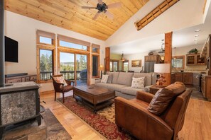 A spacious living room with a fireplace, a large sofa, and a coffee table. There is a kitchen area with wooden cabinets and a dining table.