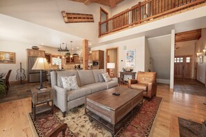 A spacious living room with a high ceiling, wooden beams, a large sofa, a coffee table, and a dining area with a chandelier.
