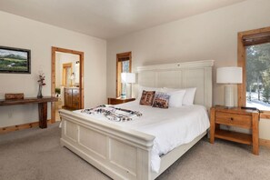Un dormitorio con una cama grande, mesitas de noche de madera, una mesilla auxiliar, un espejo y una ventana con vistas a árboles.