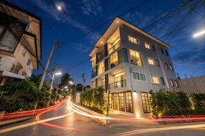 A multi-story building with balconies, a well-lit entrance, and a street with light trails indicating traffic. - Mandalar Arch 39 Hotel (Chiang Mai)