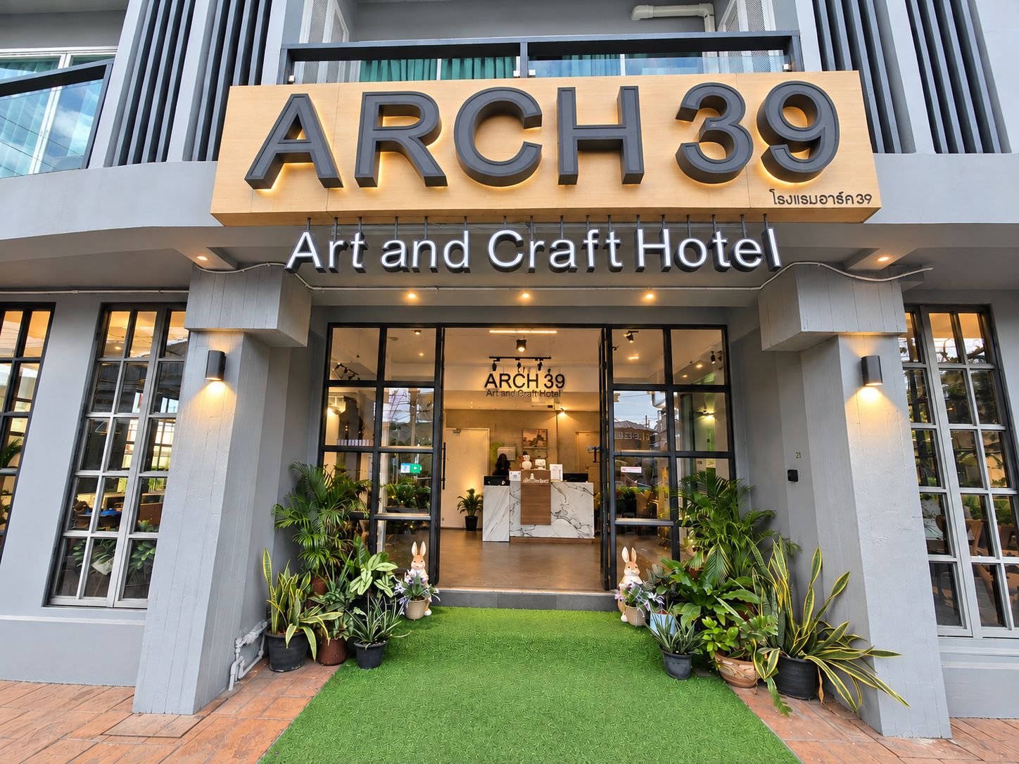 Exterior do ARCH 39 Art and Craft Hotel, com entrada visível, plantas em vasos e gramado sintético verde.