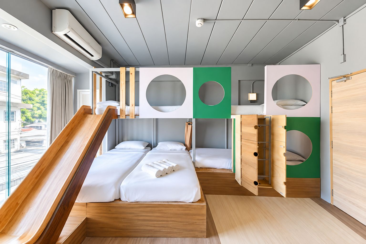 Quarto de hotel moderno com piso de madeira, cama com roupa de cama branca, um pequeno espaço de estar e uma janela com vista para o exterior.