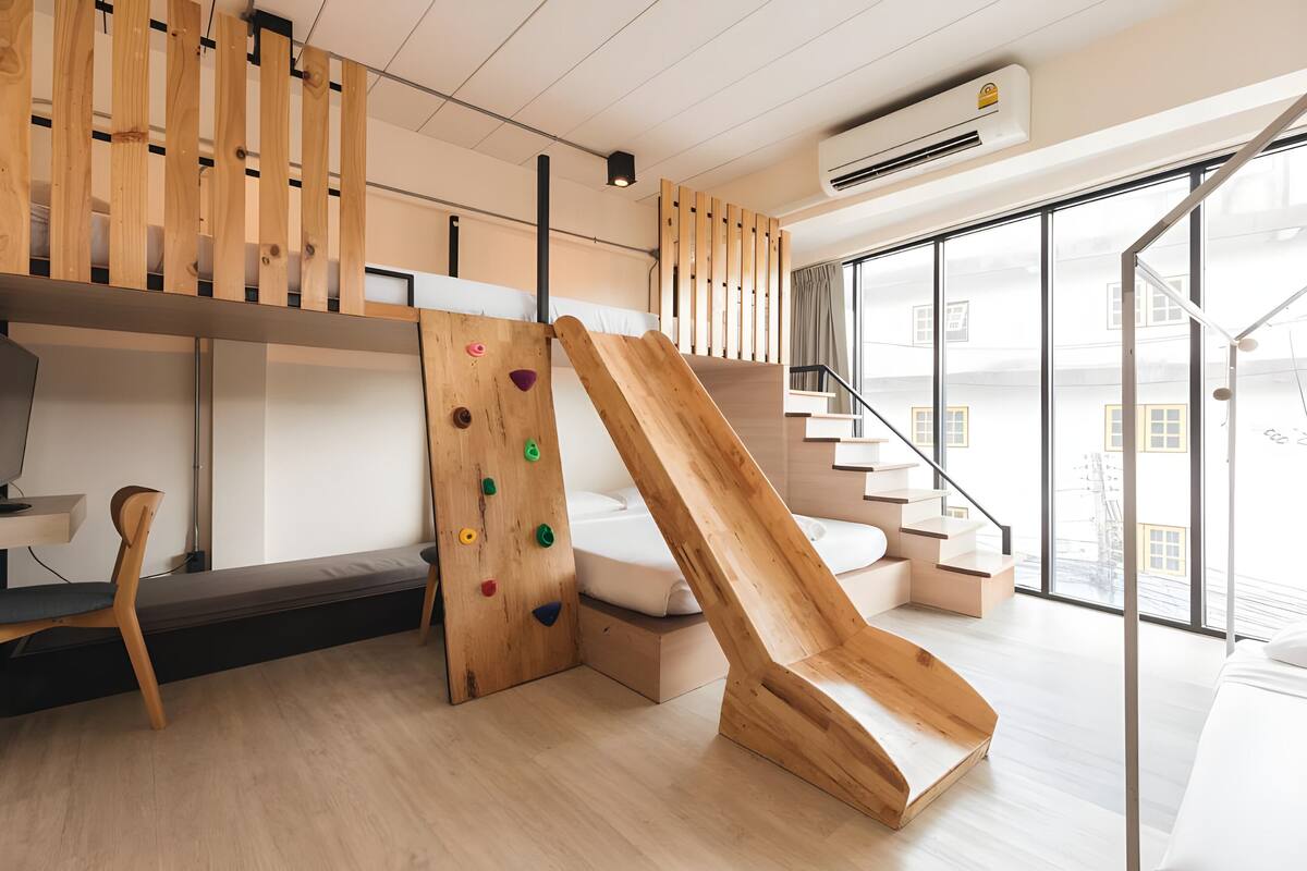 A modern room with a wooden climbing wall, a bed, a desk, and a large window with a view of buildings.