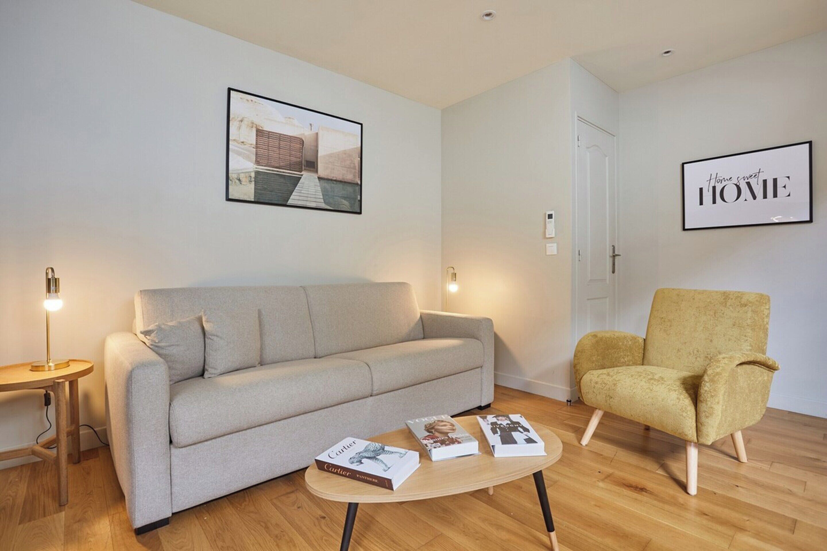 A modern living room with a grey sofa, a yellow armchair, and a coffee table with books. There are two wall-mounted lights and a framed picture above the sofa.