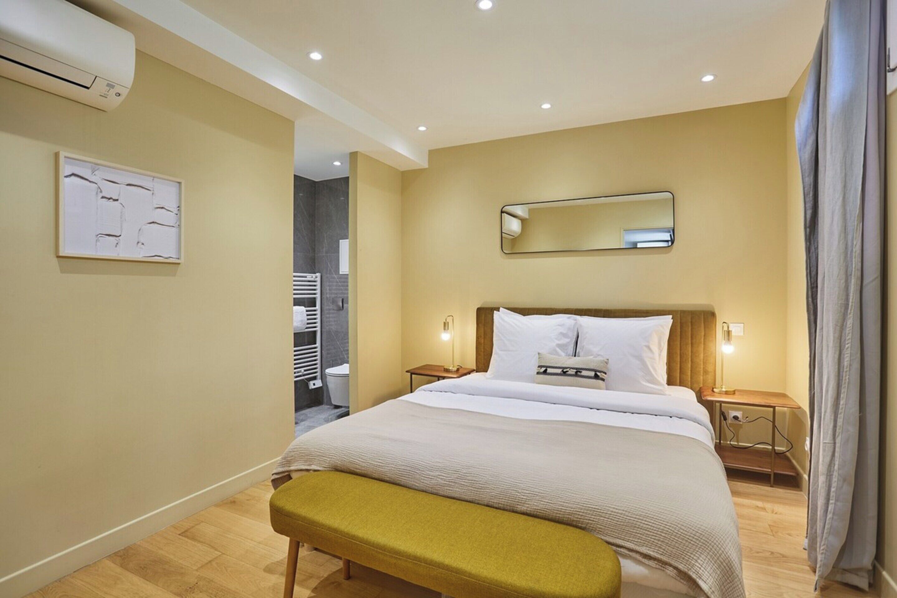 Ein Hotelzimmer mit Bett, Nachttischen, einer Bank, einem Wandspiegel und einer Klimaanlage.