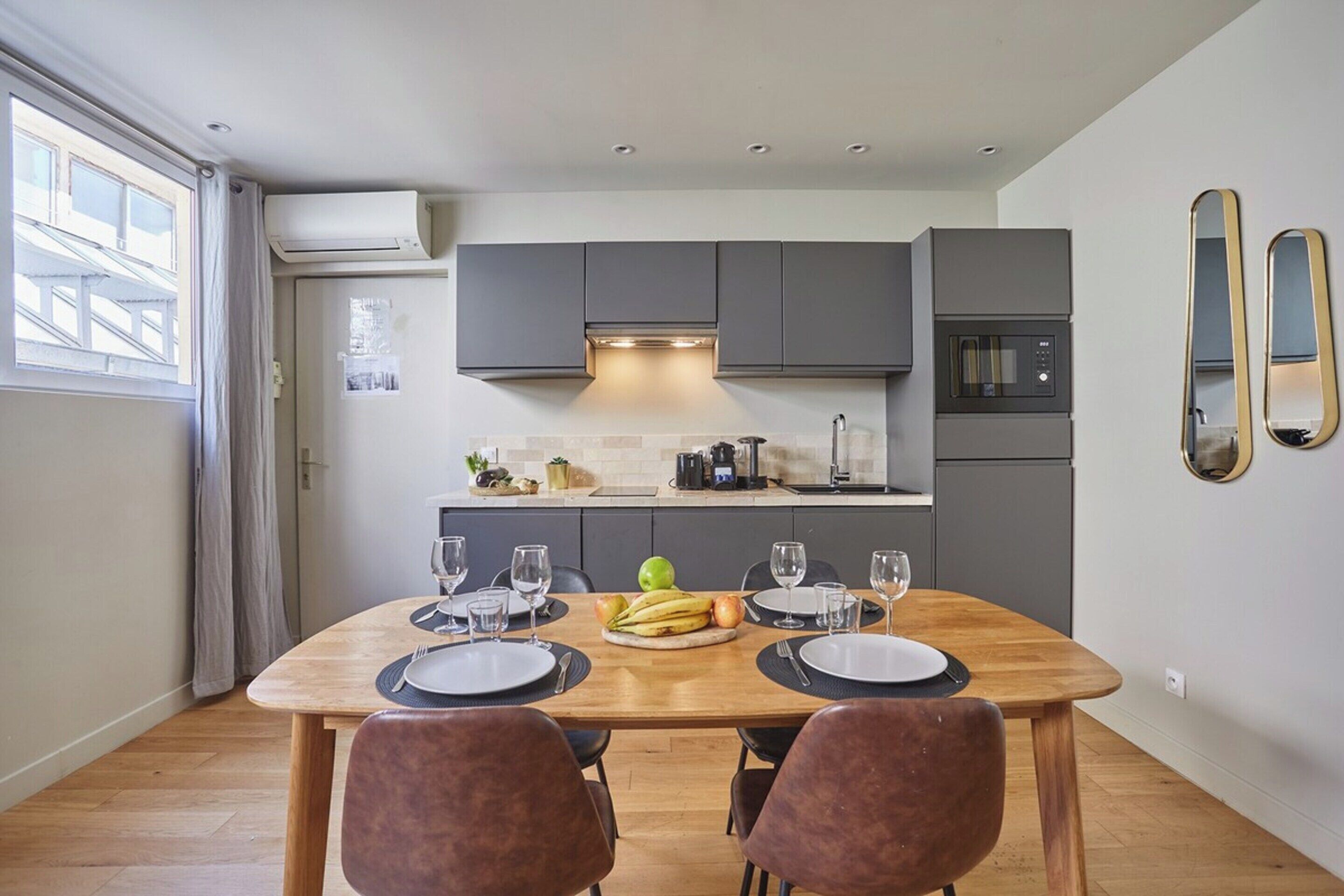 A modern kitchen with grey cabinets, a microwave, and a dining table set for four with plates, cutlery, and a fruit bowl.