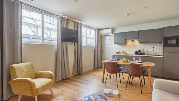 En moderne stue med sofa, spisebord med stole, et køkkenområde med skabe og et fjernsyn, samt store vinduer med gardiner.