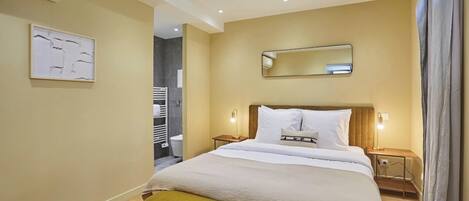 A hotel room with a bed, bedside tables, a bench, a wall-mounted mirror, and an air conditioning unit.